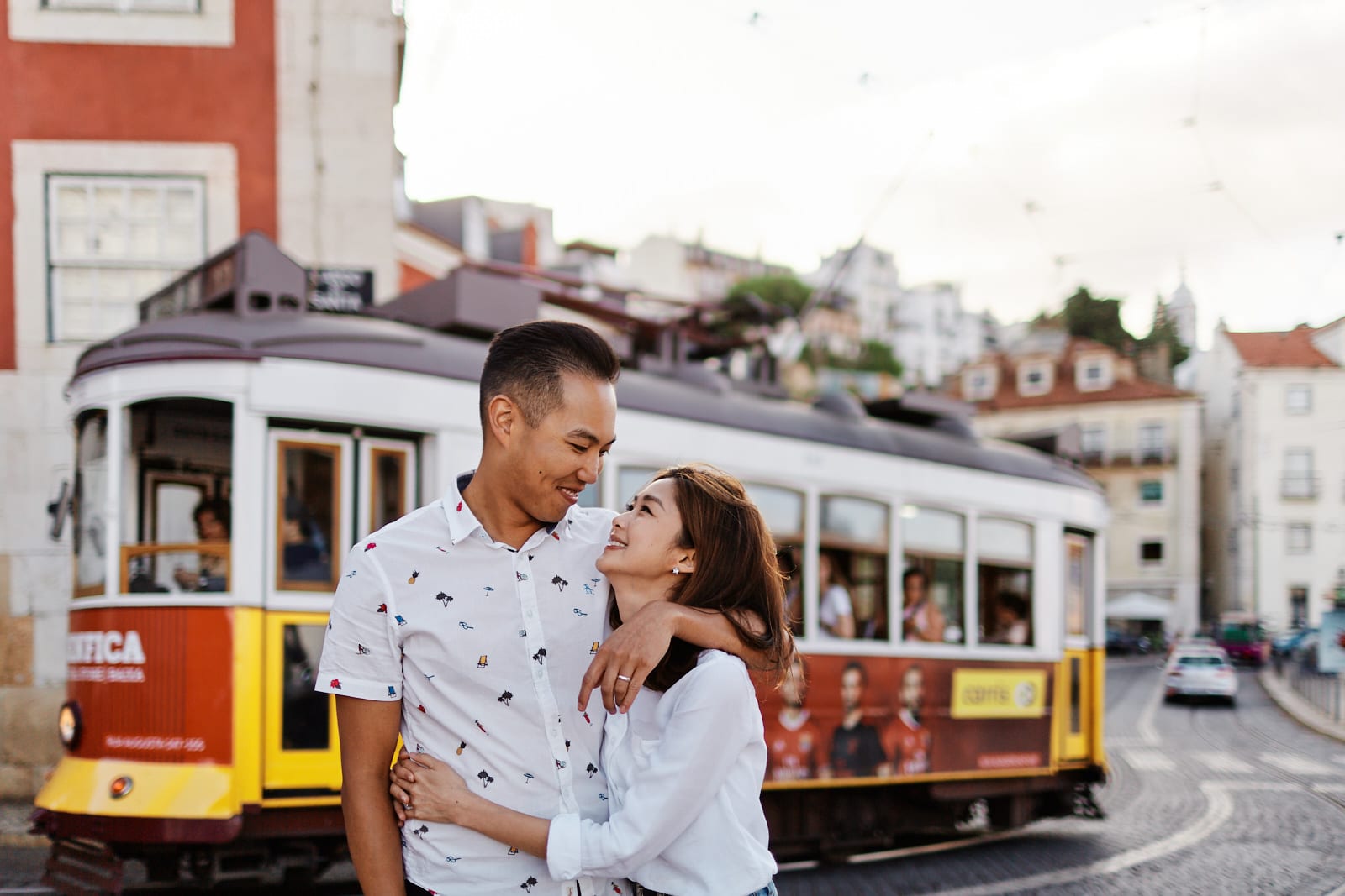 Eliza Sam and Joshua Ngo hug in the street in Lisbon, Portugal as a tram car passes behind them
