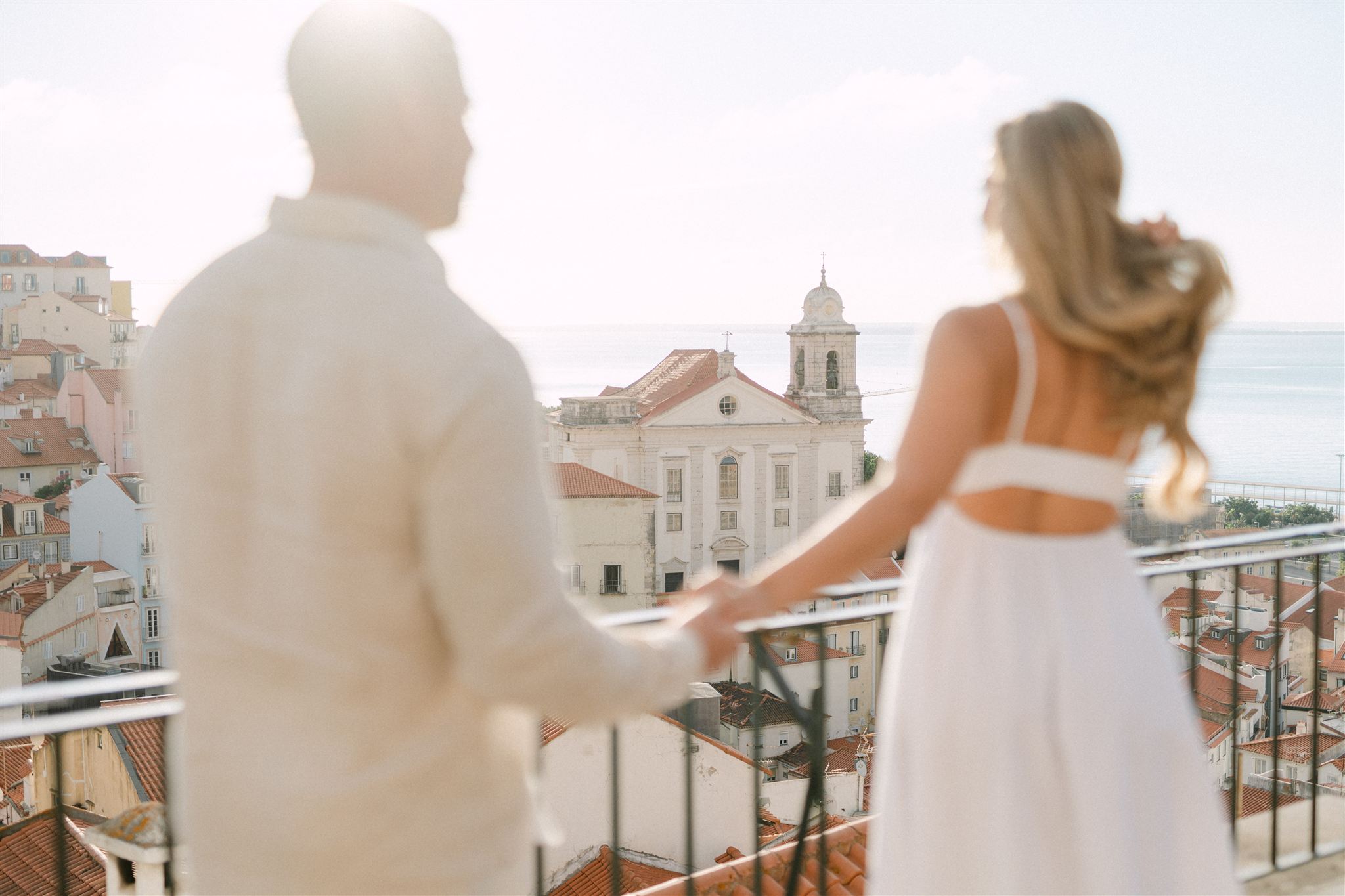 A couple hold hands while enjoying the view at Alfama's viewpoint in early morning light, during a couple photoshoot