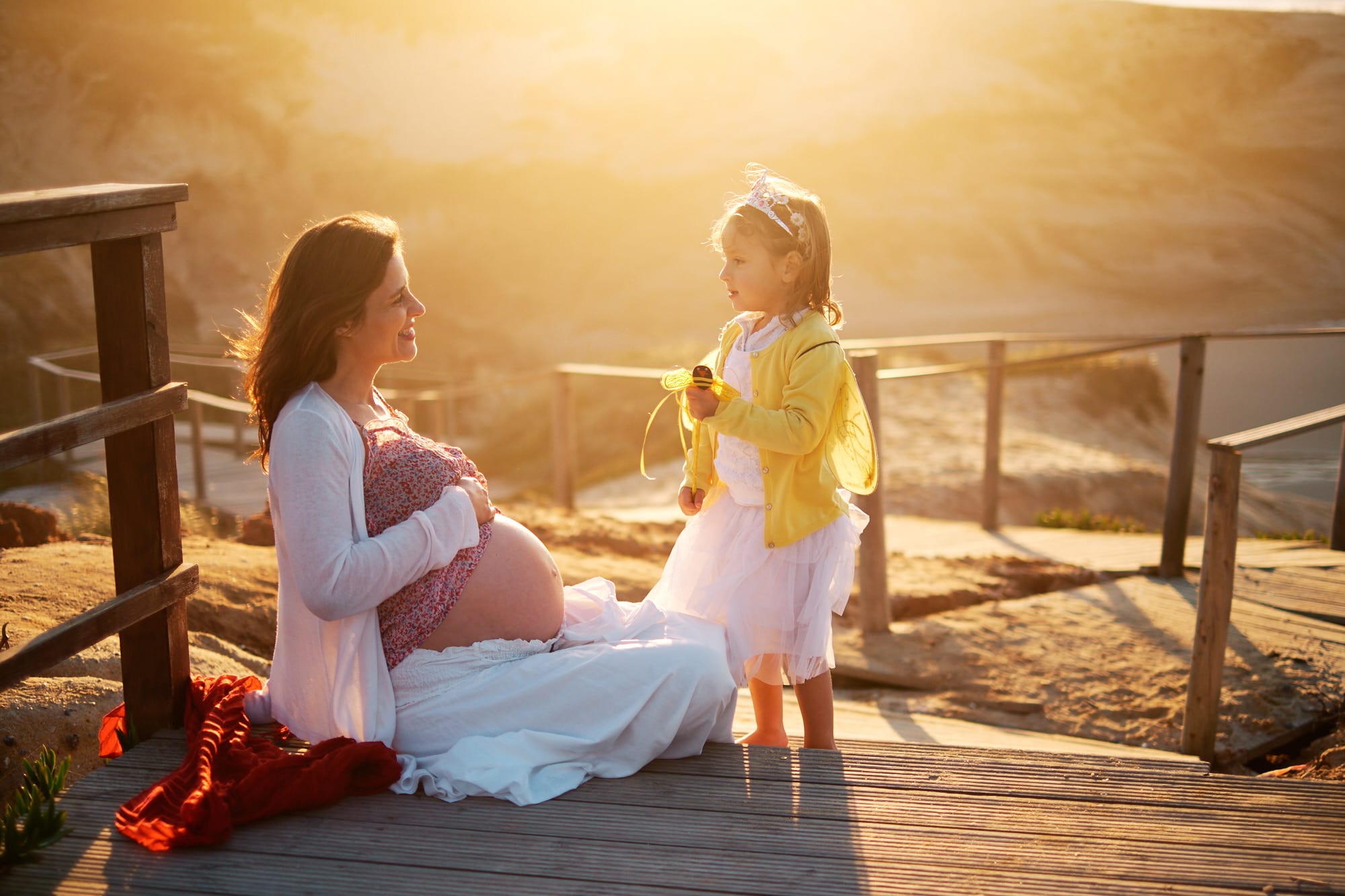 mother and daughter smile at each other in the golden light of the late afternoon during a pregnancy shoot on the beach in Portugal
